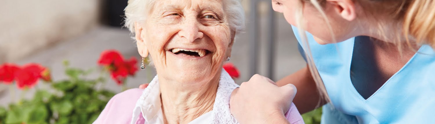 Eine lachende ältere Dame mit weißem Haar in rosa Strickjacke sitzt neben blühenden roten Blumen. Eine junge Pflegekraft in blauer Uniform beugt sich lächelnd zu ihr und berührt ihre Schulter.